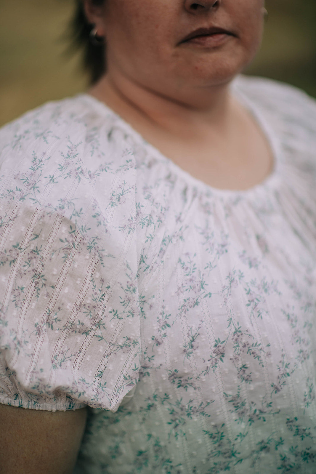 Closeup of the grace blouse, it is white with a purple and green floral pattern. The fabric has a subtle white on white polka dot and stripe texture. The neck and sleeve are both gathered.